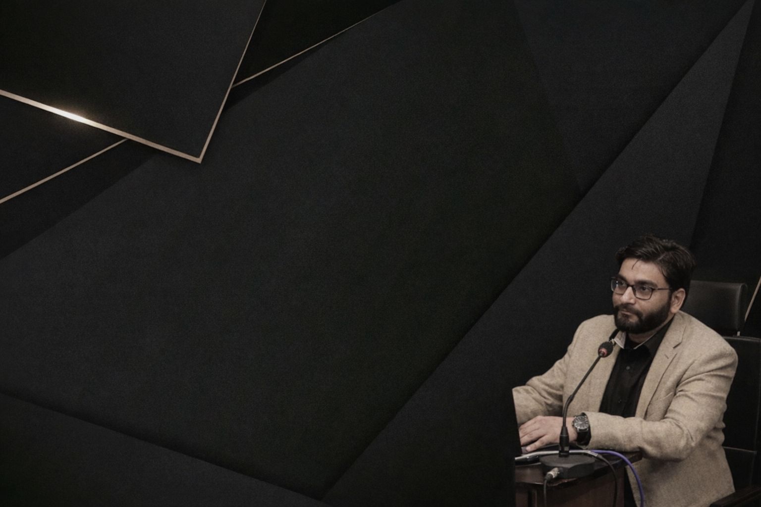 Man i kostym bakom mikrofon vid ett bord med mörk bakgrund.