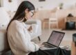 Kvinna med glasögon arbetar vid laptop med en kopp kaffe i hemmet
